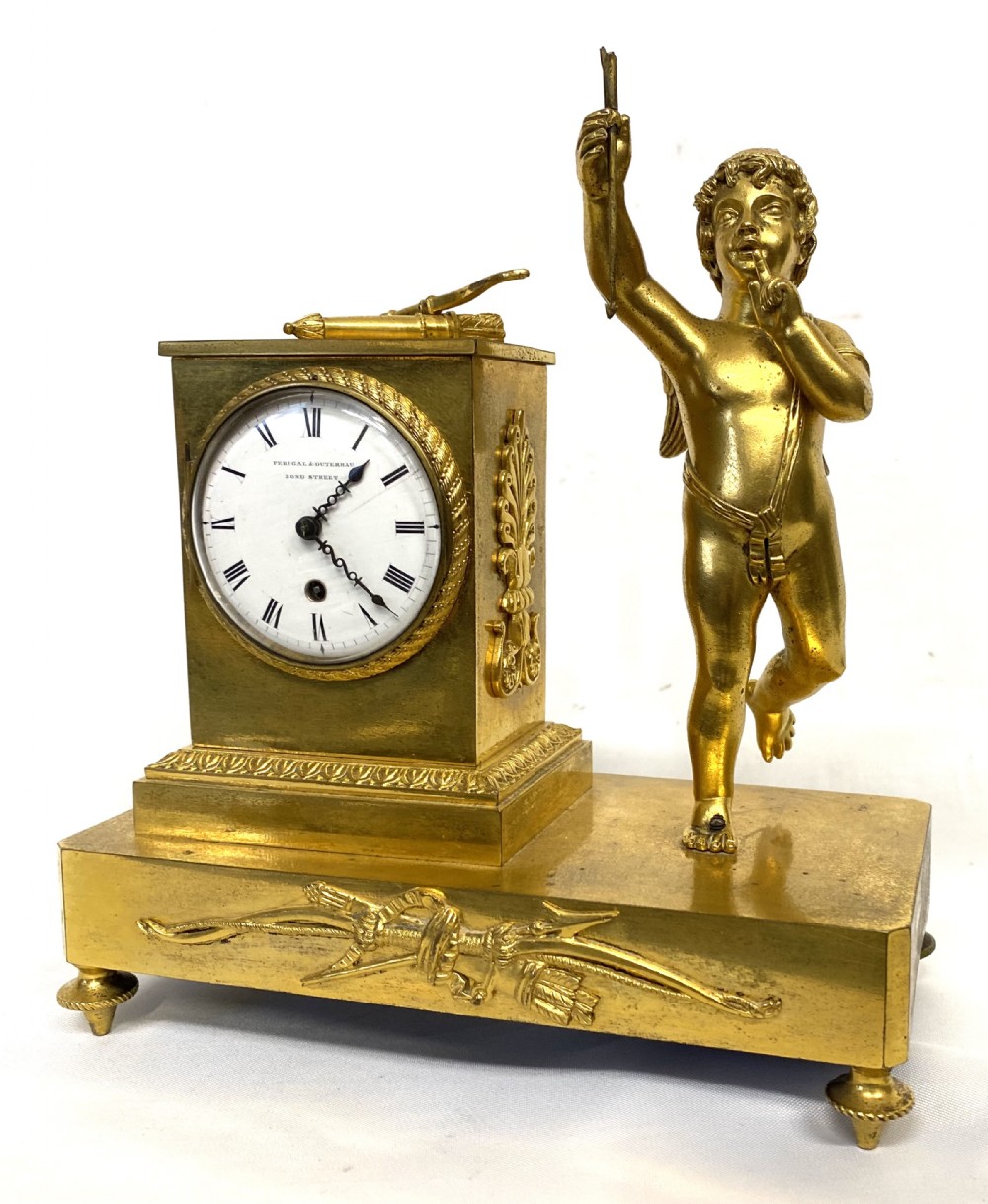An antique gilt bronze mantel clock featuring a white enamel dial alongside a standing Cupid figure holding an arrow, set upon a decorative gilded plinth base.