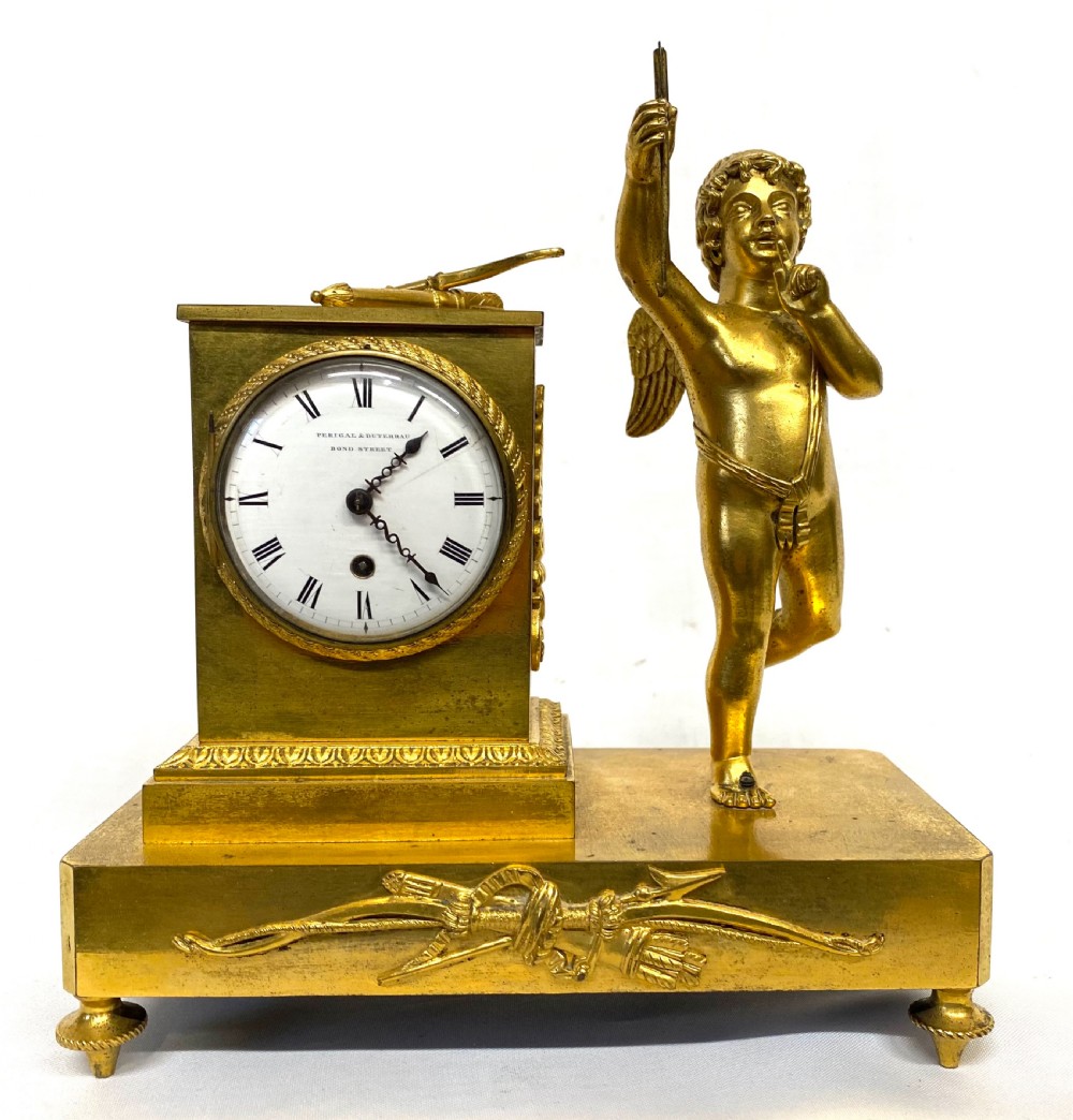 An antique gilt bronze mantel clock featuring a white enamel dial alongside a standing Cupid figure holding an arrow, set upon a decorative gilded plinth base. View IV