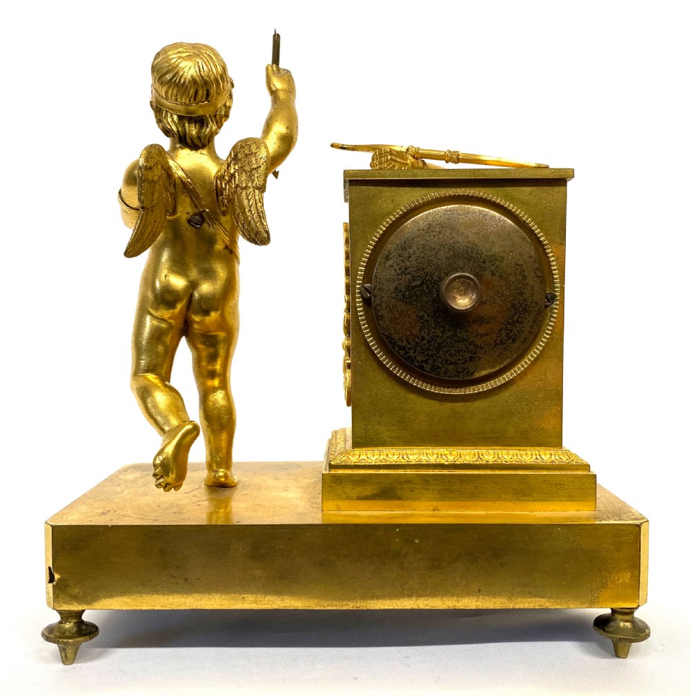 An antique gilt bronze mantel clock featuring a white enamel dial alongside a standing Cupid figure holding an arrow, set upon a decorative gilded plinth base. View IX