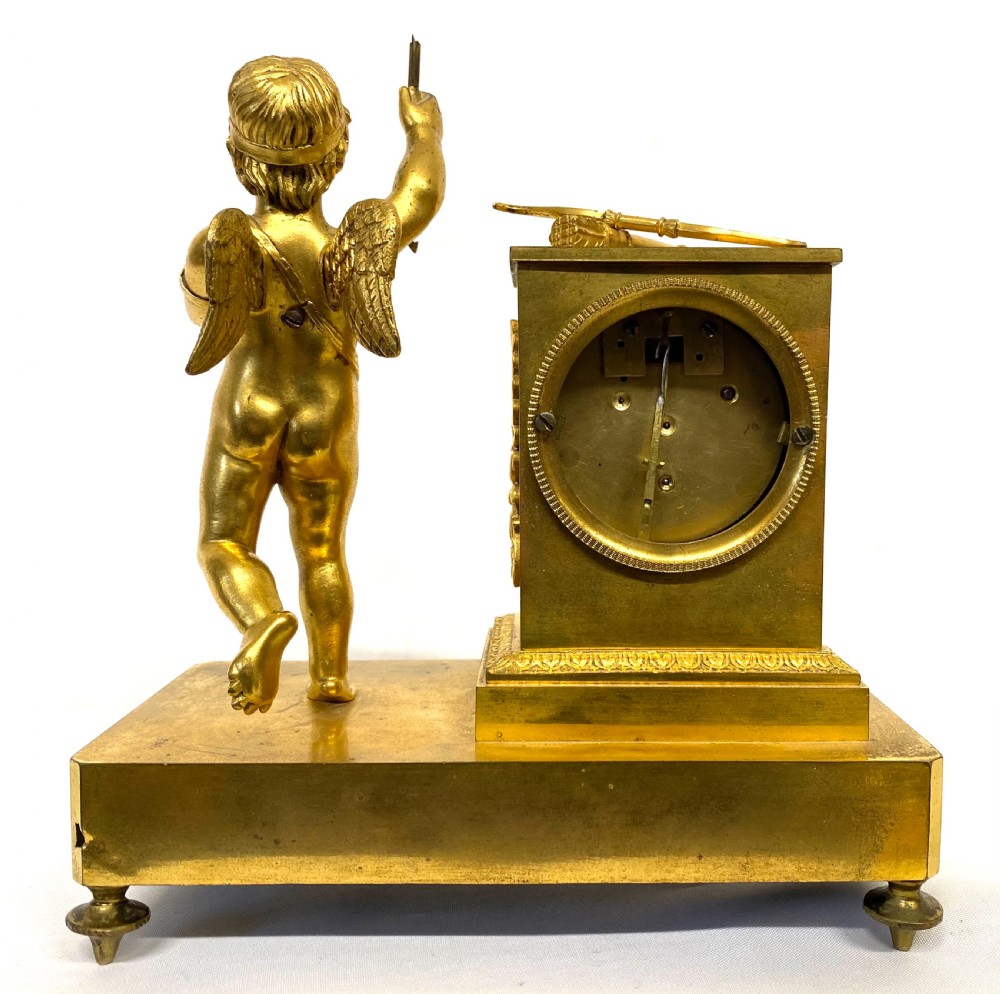 An antique gilt bronze mantel clock featuring a white enamel dial alongside a standing Cupid figure holding an arrow, set upon a decorative gilded plinth base. View X