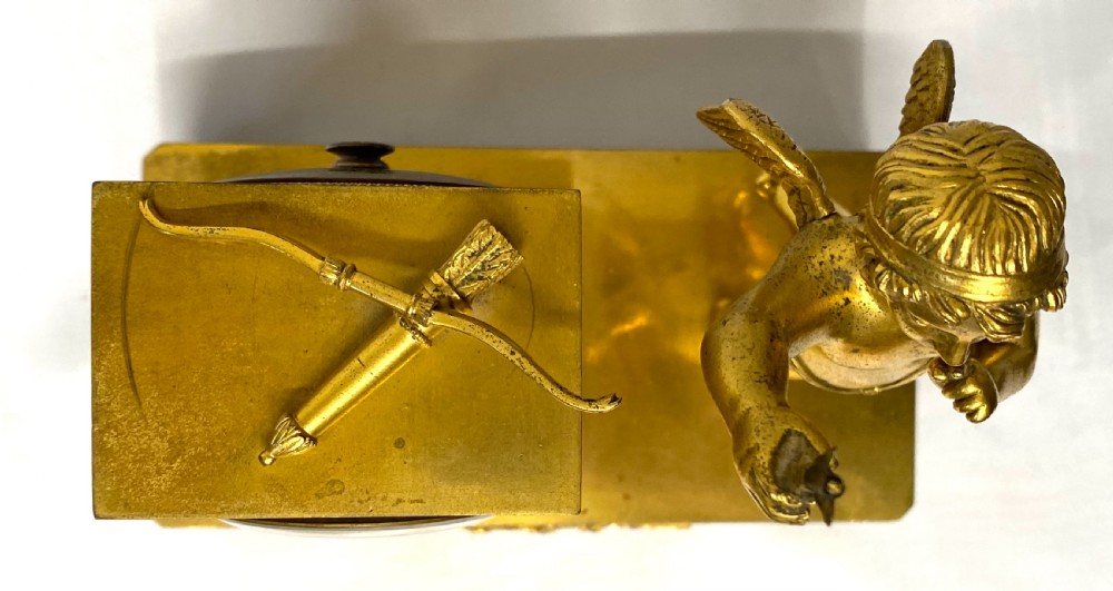 An antique gilt bronze mantel clock featuring a white enamel dial alongside a standing Cupid figure holding an arrow, set upon a decorative gilded plinth base. View XII