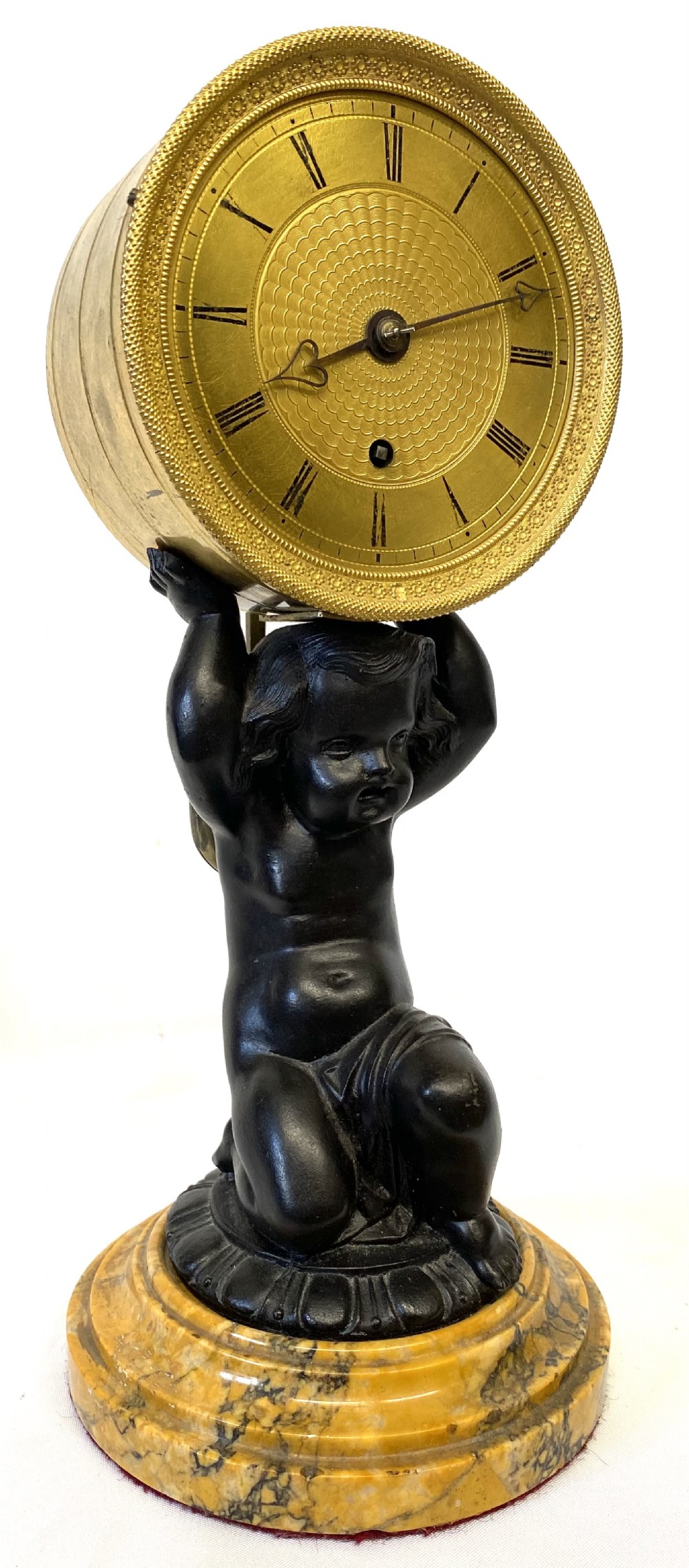 An antique gilt brass and dark patinated bronze mantel clock depicting a kneeling cherub supporting a circular drum dial above a veined yellow marble base.