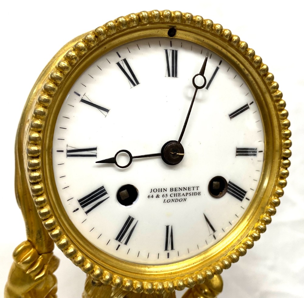 A gilt bronze Victorian mantel clock featuring a sculpted cherub supporting a circular white enamel clock dial signed John Bennett London. View VIII