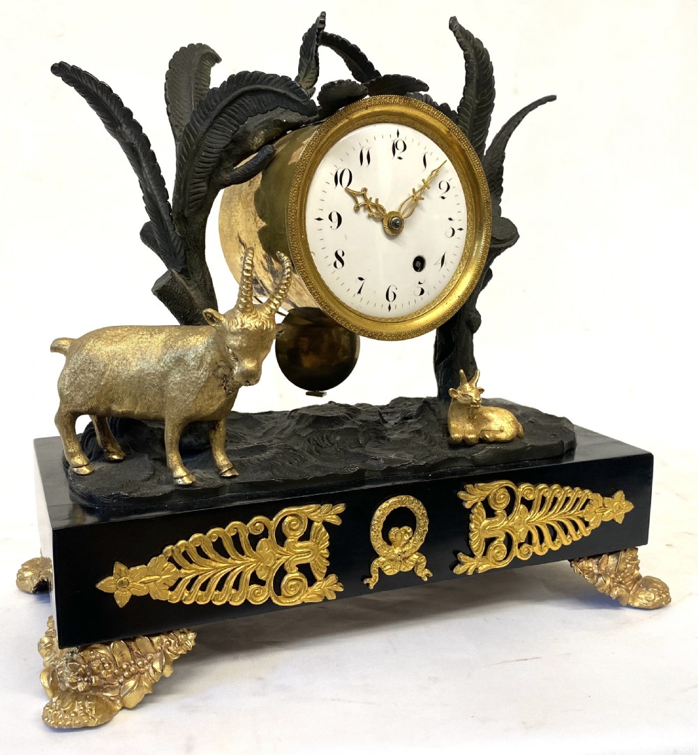 An antique Regency gilt and patinated bronze mantel clock featuring sculpted goats beneath palm trees on a black rectangular base with scrolled gilt feet.
