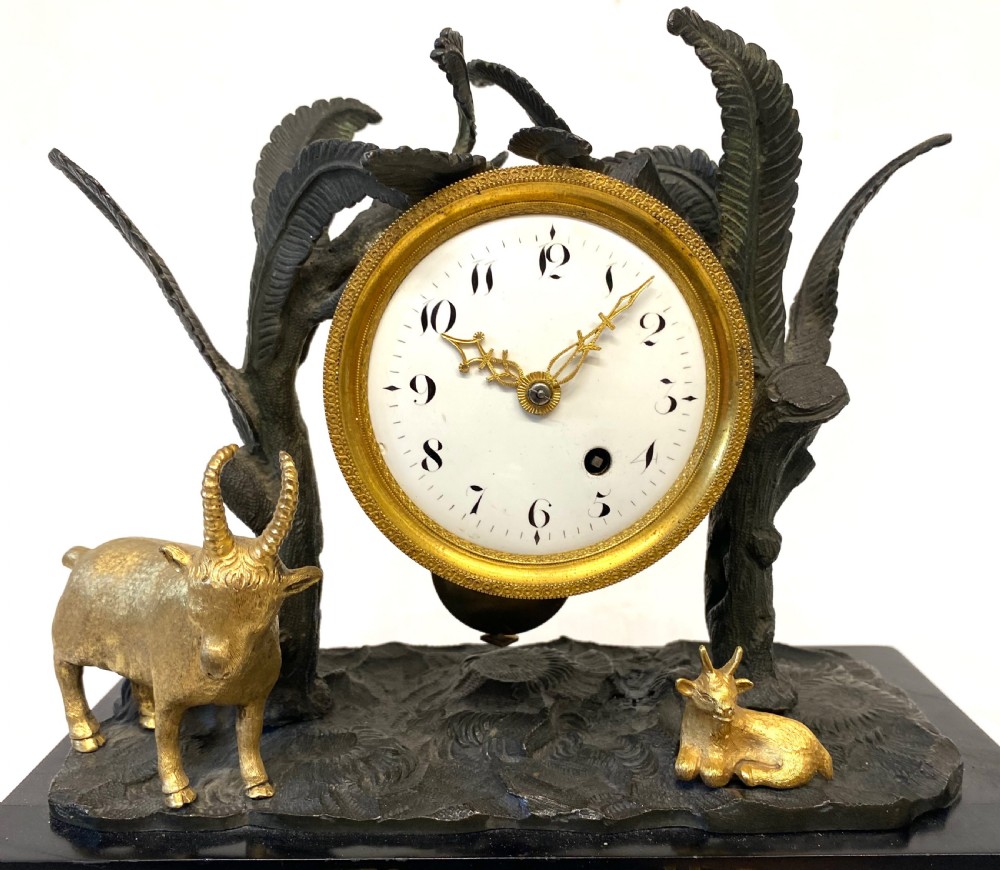 An antique Regency gilt and patinated bronze mantel clock featuring sculpted goats beneath palm trees on a black rectangular base with scrolled gilt feet. View II