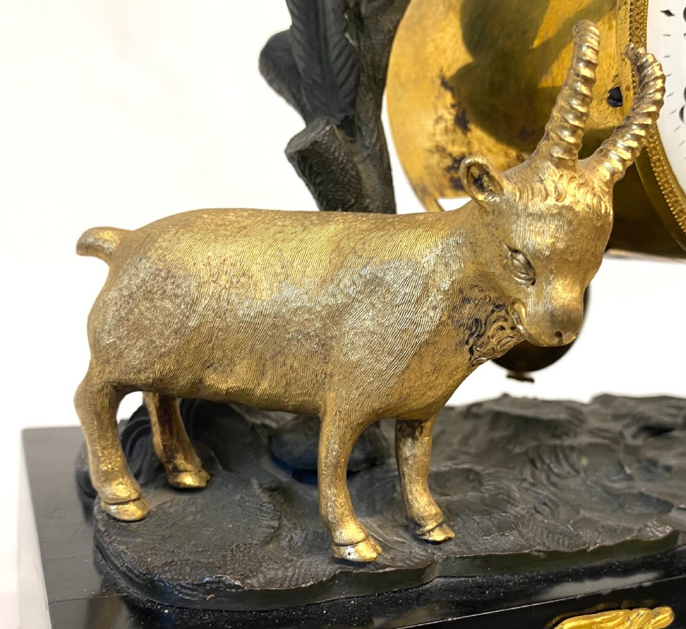 An antique Regency gilt and patinated bronze mantel clock featuring sculpted goats beneath palm trees on a black rectangular base with scrolled gilt feet. View V