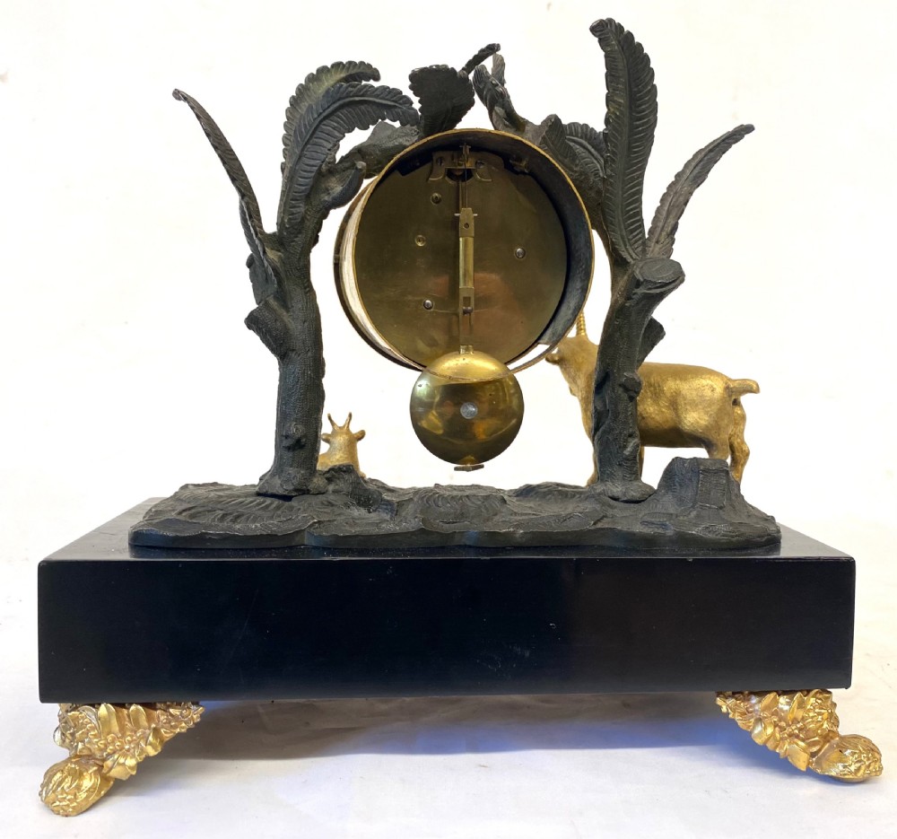 An antique Regency gilt and patinated bronze mantel clock featuring sculpted goats beneath palm trees on a black rectangular base with scrolled gilt feet. View IX