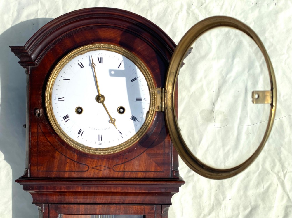 An antique flame mahogany wall clock featuring a round white enamel dial, brass bezel, and a glazed door displaying a gridiron pendulum. View V