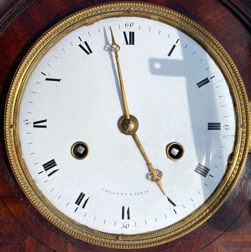 An antique flame mahogany wall clock featuring a round white enamel dial, brass bezel, and a glazed door displaying a gridiron pendulum. View VI