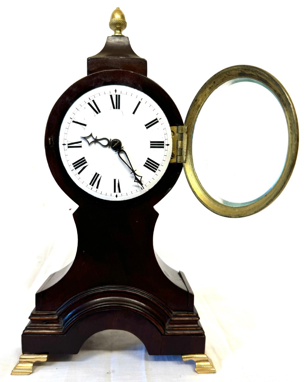 A dark mahogany miniature balloon clock featuring a circular white enamel dial with Roman numerals, a brass acorn finial, and brass feet. View IV