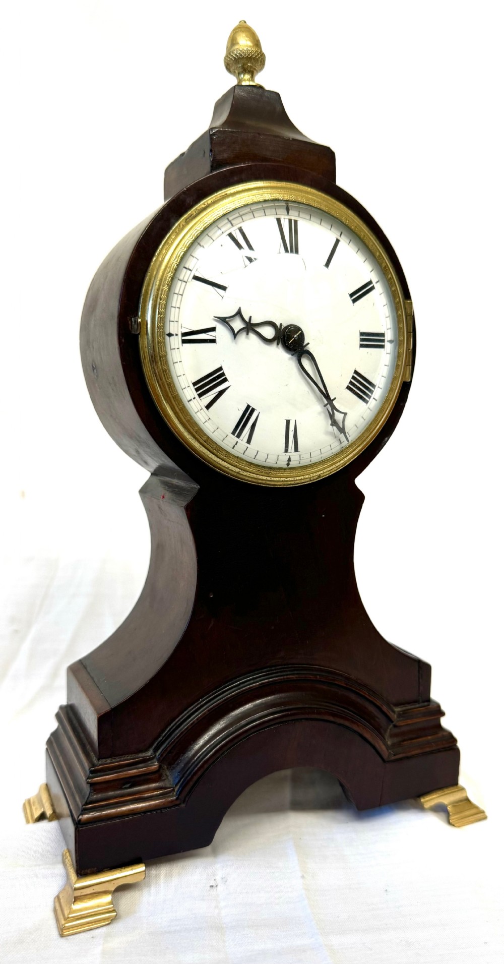 A dark mahogany miniature balloon clock featuring a circular white enamel dial with Roman numerals, a brass acorn finial, and brass feet. View VI