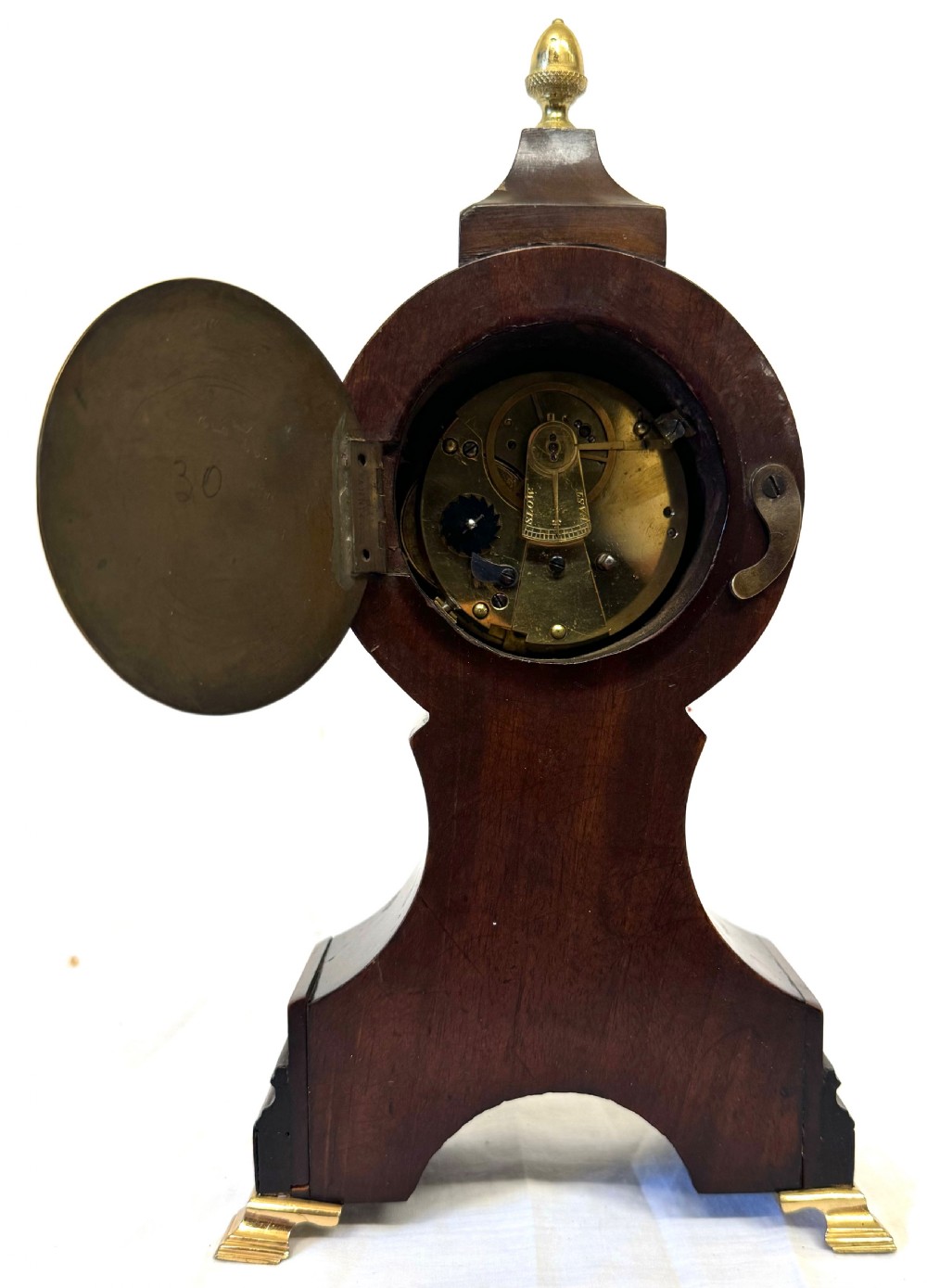 A dark mahogany miniature balloon clock featuring a circular white enamel dial with Roman numerals, a brass acorn finial, and brass feet. View IX