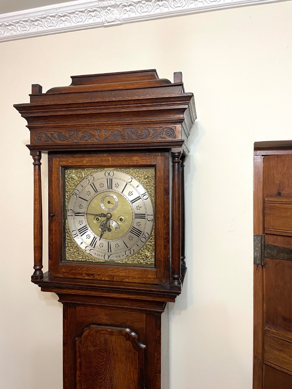 An antique dark oak longcase grandfather clock featuring a carved wooden hood, brass dial with Roman numerals, and a tall rectangular trunk door. View IV