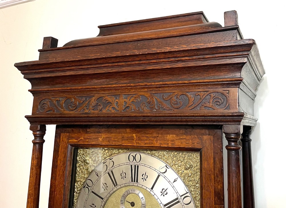 An antique dark oak longcase grandfather clock featuring a carved wooden hood, brass dial with Roman numerals, and a tall rectangular trunk door. View XXIV