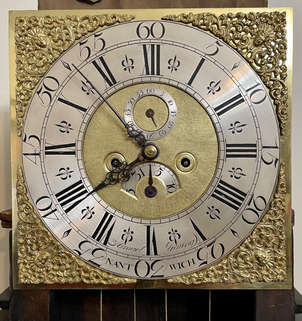 An antique dark oak longcase grandfather clock featuring a carved wooden hood, brass dial with Roman numerals, and a tall rectangular trunk door. View XXVI