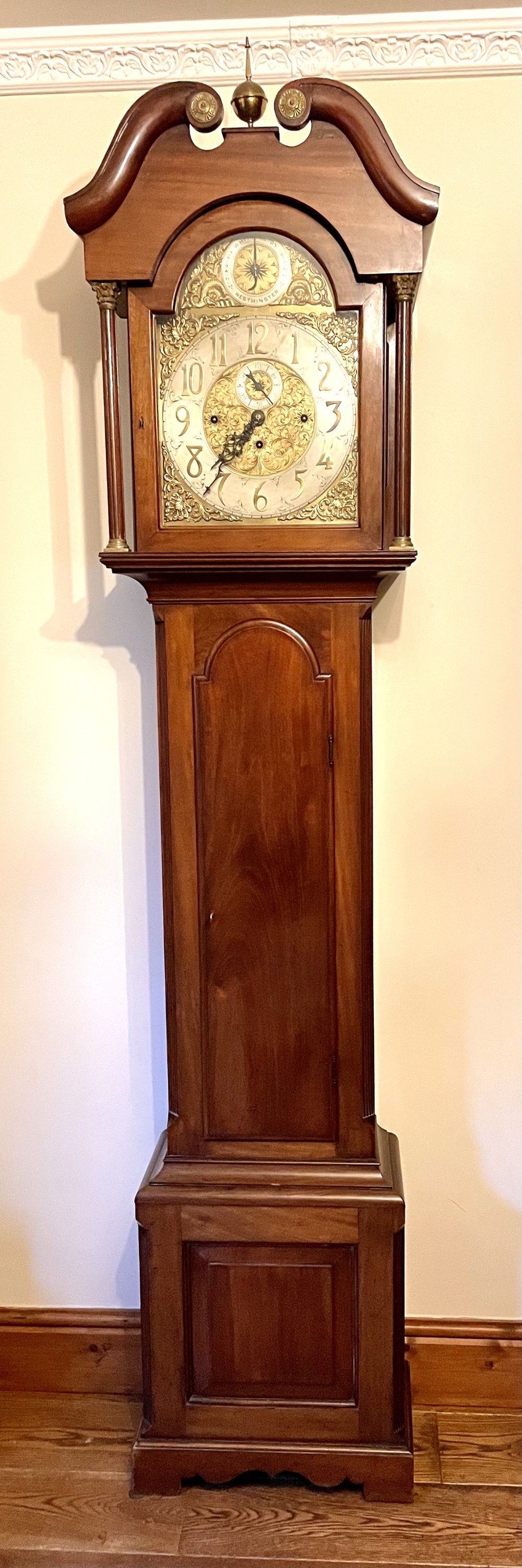 A mahogany longcase clock featuring a swan-neck pediment, brass arch dial with silvered chapter ring, fluted columns, and a shaped trunk door.