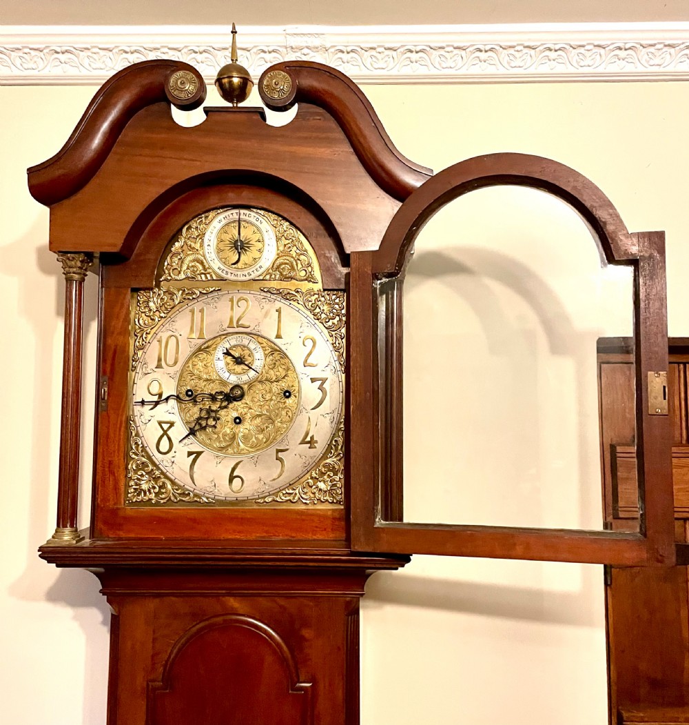 A mahogany longcase clock featuring a swan-neck pediment, brass arch dial with silvered chapter ring, fluted columns, and a shaped trunk door. View VII