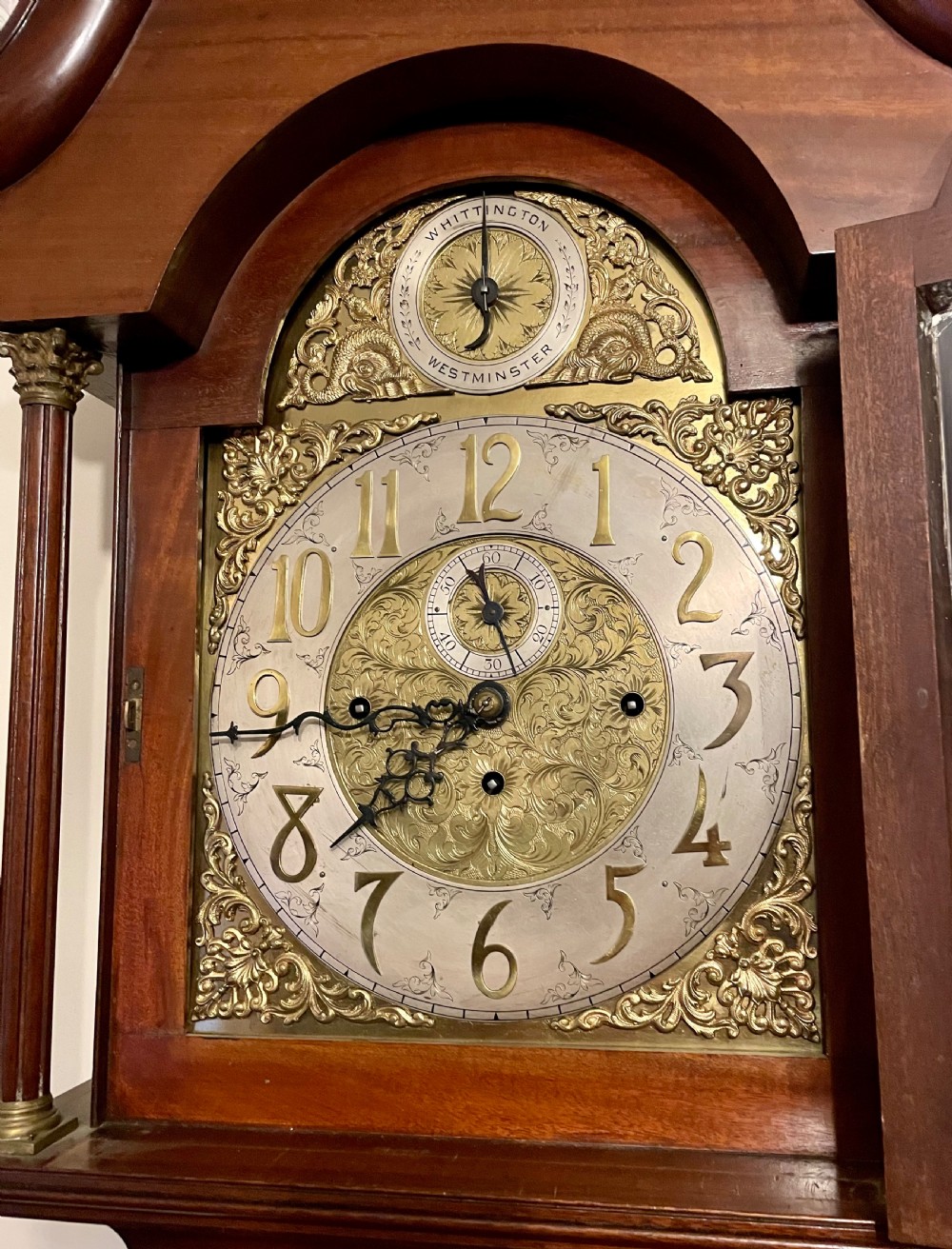 A mahogany longcase clock featuring a swan-neck pediment, brass arch dial with silvered chapter ring, fluted columns, and a shaped trunk door. View VIII
