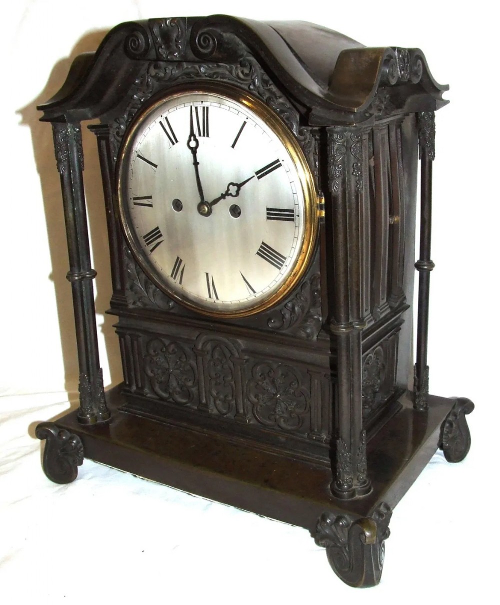 A dark brown patinated bronze mantel clock featuring Gothic architectural columns, decorative relief carvings, scroll feet, and a silvered circular dial with Roman numerals.