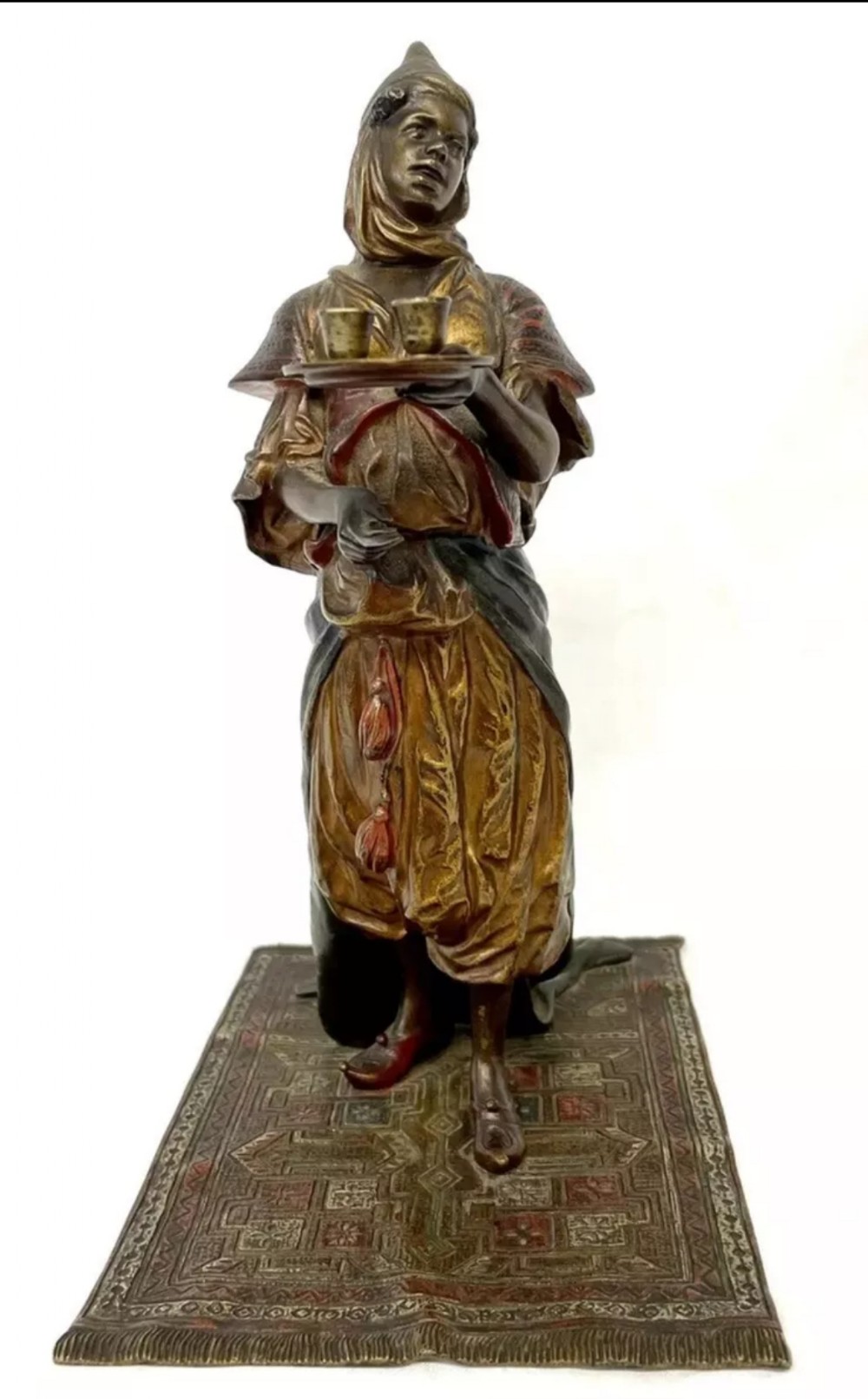 An antique cold-painted bronze statuette depicting an Arab woman carrying a tea tray while standing on an engraved rug base.