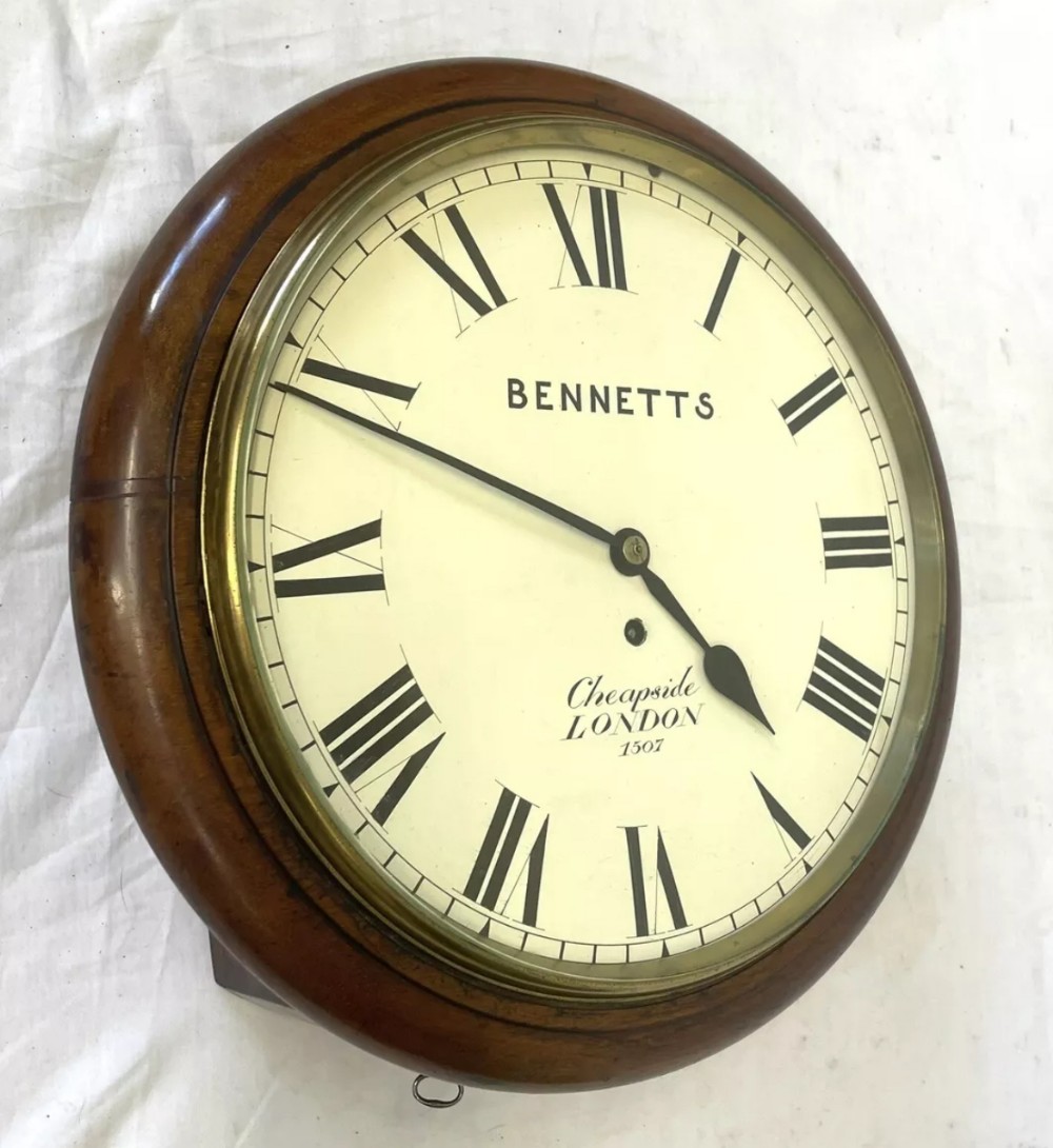 An antique circular mahogany wall clock featuring an off-white dial with black Roman numerals, brass bezel, and Bennetts Cheapside London inscription.