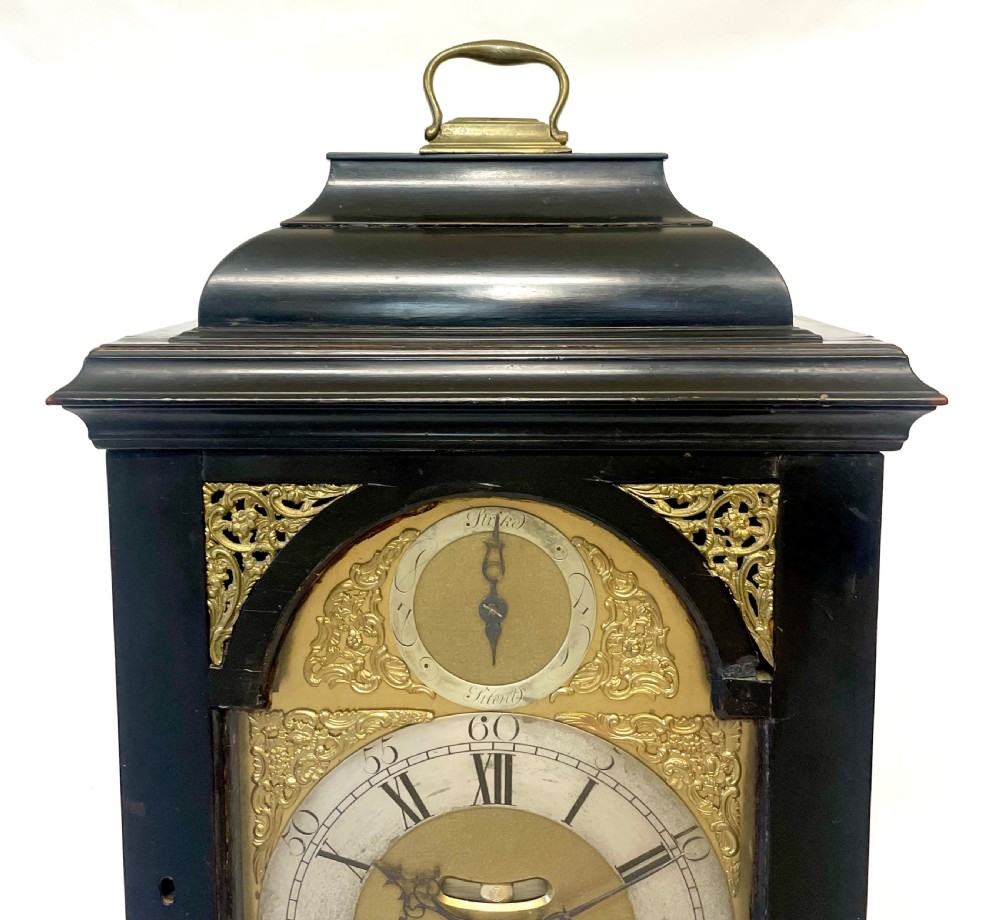 An 18th-century ebonised wooden bracket clock featuring a brass dial with silvered chapter rings, decorative corner spandrels, a strike/silent sub-dial, and a brass carrying handle on top. View V