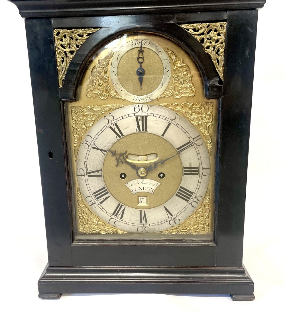 An 18th-century ebonised wooden bracket clock featuring a brass dial with silvered chapter rings, decorative corner spandrels, a strike/silent sub-dial, and a brass carrying handle on top. View VI