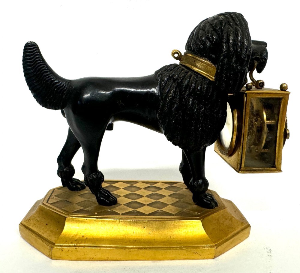 A French gilt and patinated bronze timepiece featuring a standing dog figure holding a square glass clock case in its mouth on a gilded base. View V