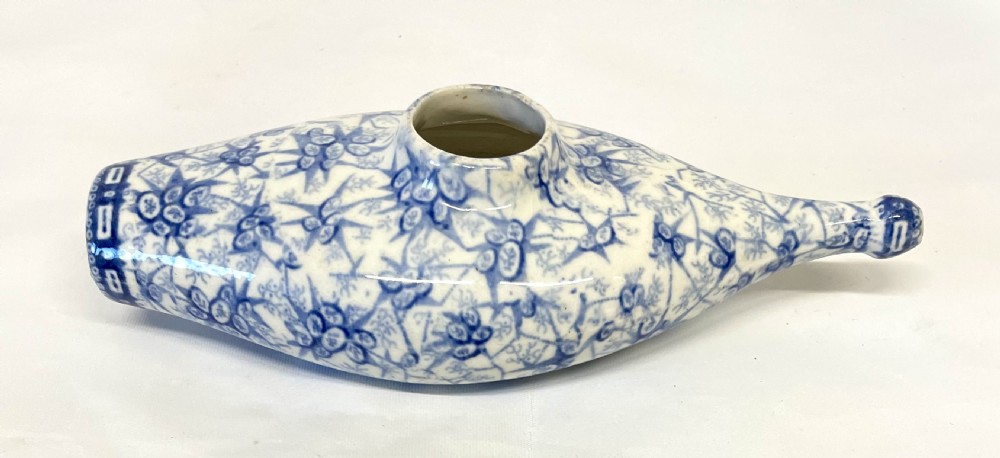A nineteenth-century ceramic baby feeding bottle decorated with blue and white floral transferware motifs, featuring a central circular opening and an elongated spout.