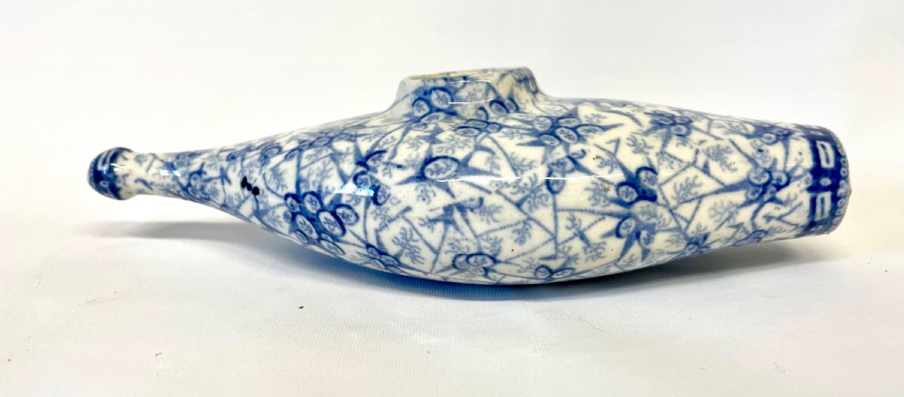 A nineteenth-century ceramic baby feeding bottle decorated with blue and white floral transferware motifs, featuring a central circular opening and an elongated spout. View IV
