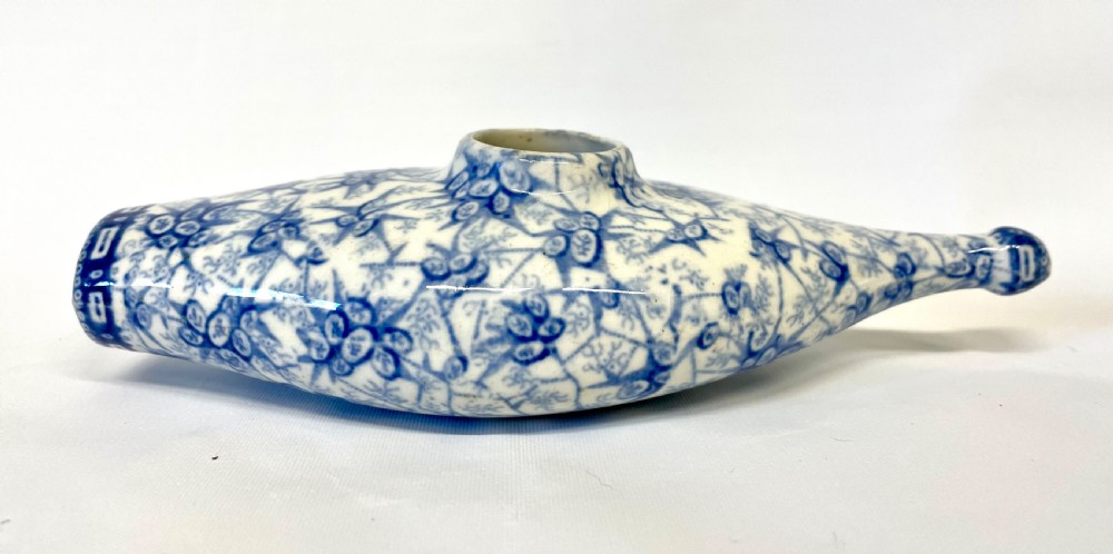 A nineteenth-century ceramic baby feeding bottle decorated with blue and white floral transferware motifs, featuring a central circular opening and an elongated spout. View V