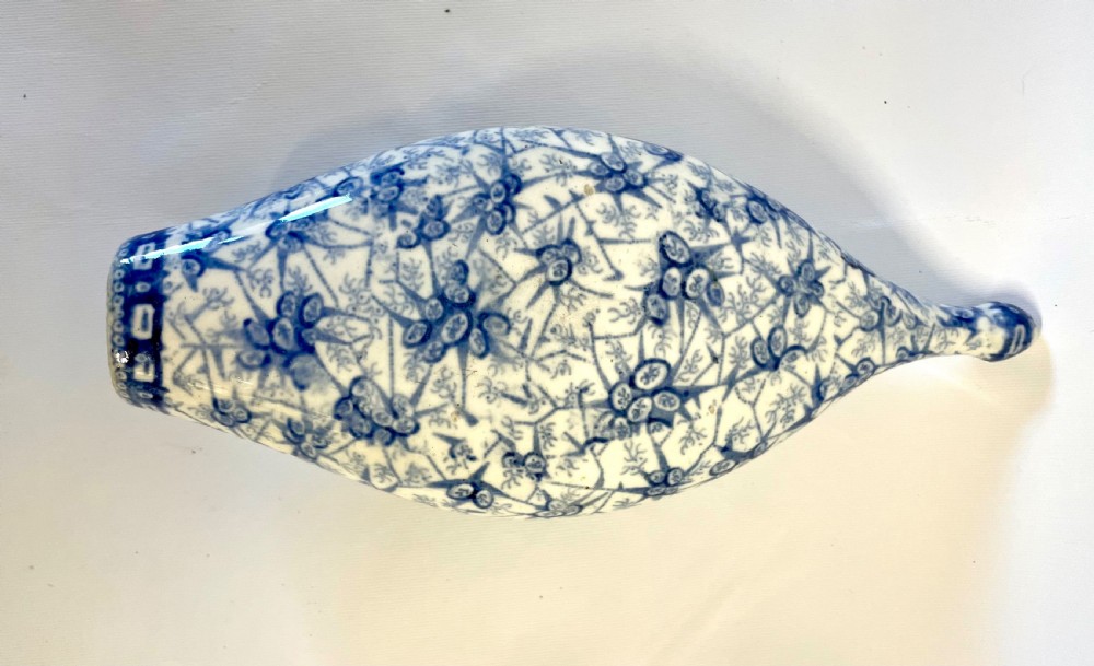 A nineteenth-century ceramic baby feeding bottle decorated with blue and white floral transferware motifs, featuring a central circular opening and an elongated spout. View IX