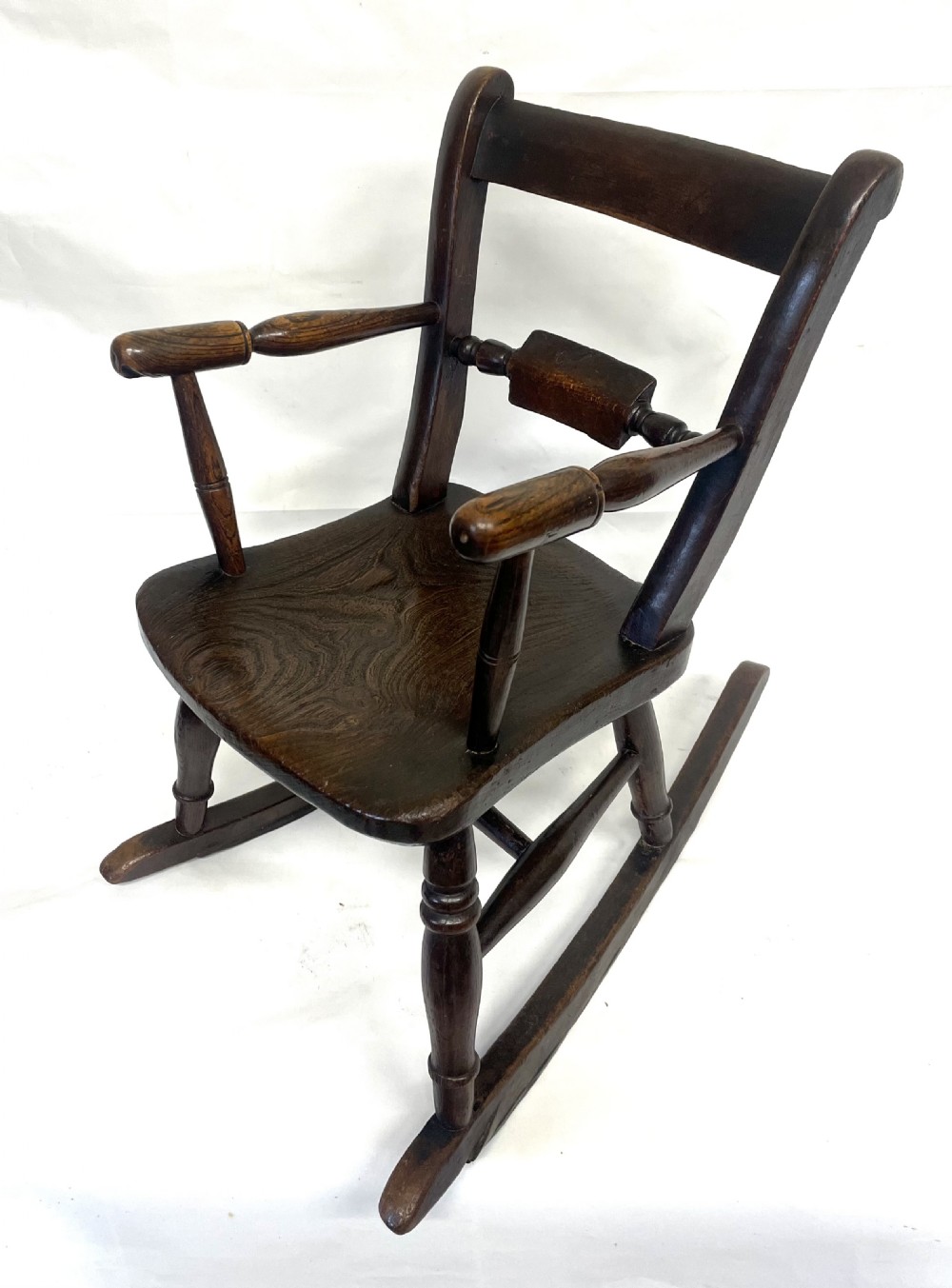 A dark brown wooden childs or dolls rocking armchair featuring turned legs, curved armrests, and smooth wooden rockers. View II