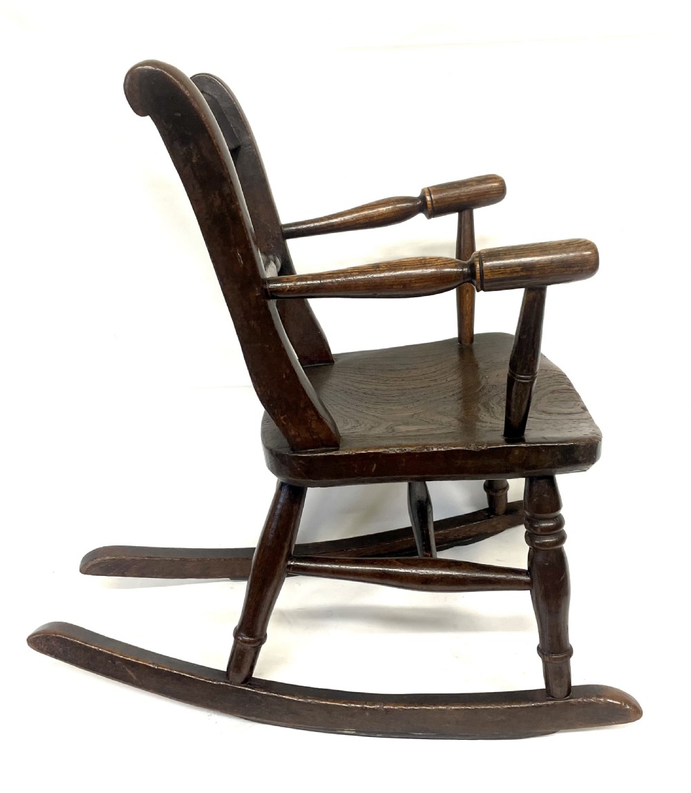 A dark brown wooden childs or dolls rocking armchair featuring turned legs, curved armrests, and smooth wooden rockers. View X