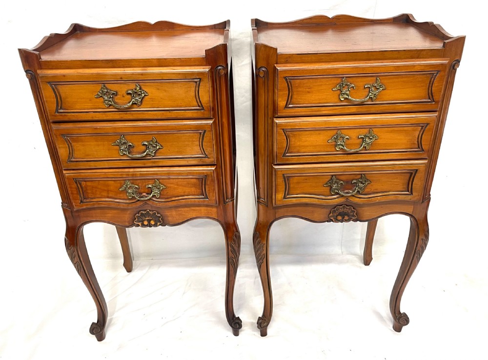 A pair of French satin walnut bedside cabinets featuring three drawers with brass handles, carved shell details on the apron, and slender cabriole legs.