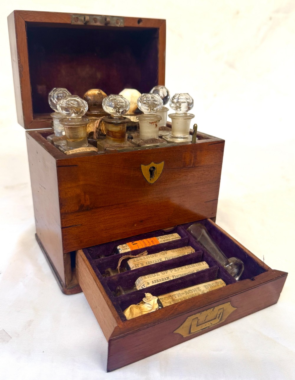 An antique wooden apothecary box shown open with glass stoppered bottles in top compartments and labelled vials in a velvet-lined lower drawer.
