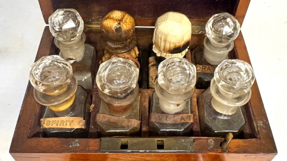 An antique wooden apothecary box shown open with glass stoppered bottles in top compartments and labelled vials in a velvet-lined lower drawer. View IV