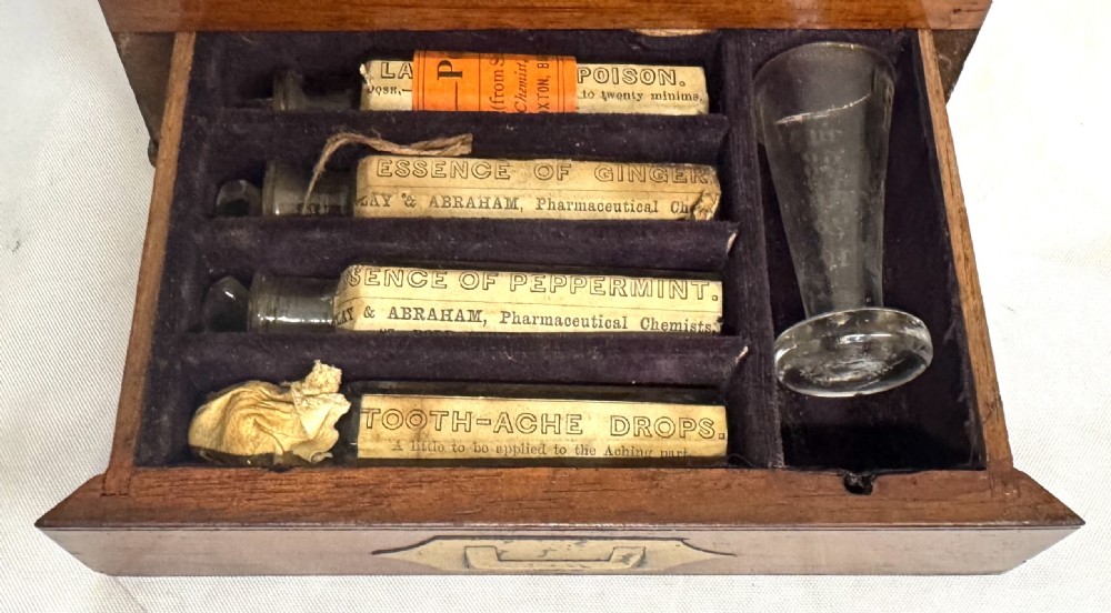 An antique wooden apothecary box shown open with glass stoppered bottles in top compartments and labelled vials in a velvet-lined lower drawer. View V