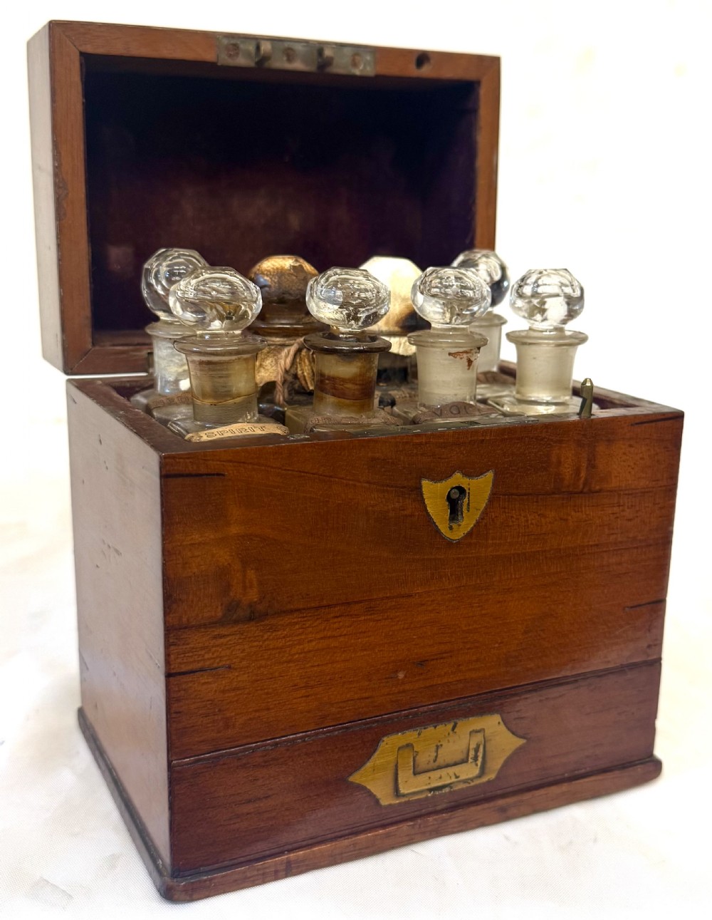 An antique wooden apothecary box shown open with glass stoppered bottles in top compartments and labelled vials in a velvet-lined lower drawer. View VI