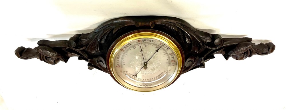 A horizontally orientated Victorian carved oak aneroid barometer featuring a central circular brass dial flanked by carved leaf motifs and bird figures. View VI