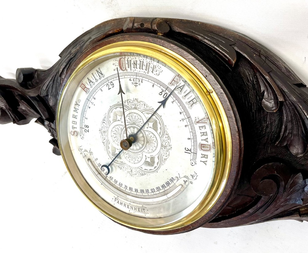 A horizontally orientated Victorian carved oak aneroid barometer featuring a central circular brass dial flanked by carved leaf motifs and bird figures. View IX