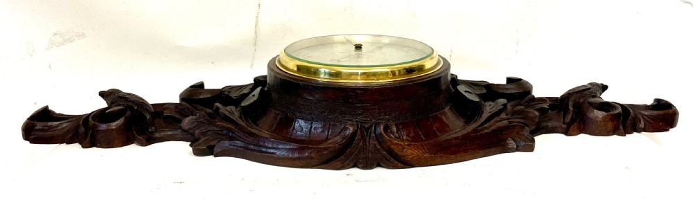 A horizontally orientated Victorian carved oak aneroid barometer featuring a central circular brass dial flanked by carved leaf motifs and bird figures. View X