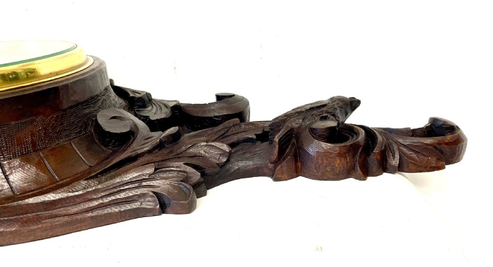 A horizontally orientated Victorian carved oak aneroid barometer featuring a central circular brass dial flanked by carved leaf motifs and bird figures. View XII