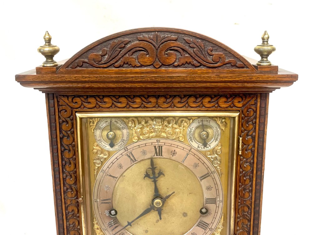 A carved oak bracket clock featuring an arched pediment, brass corner finials, side fretwork panels, and a brass dial with silvered chapter ring. View V