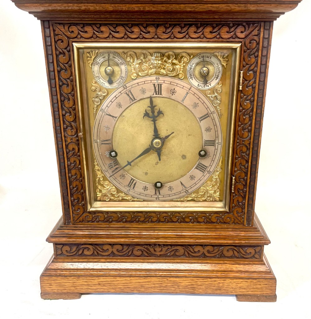 A carved oak bracket clock featuring an arched pediment, brass corner finials, side fretwork panels, and a brass dial with silvered chapter ring. View VI