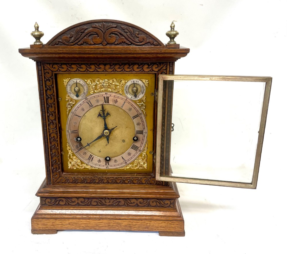 A carved oak bracket clock featuring an arched pediment, brass corner finials, side fretwork panels, and a brass dial with silvered chapter ring. View VII