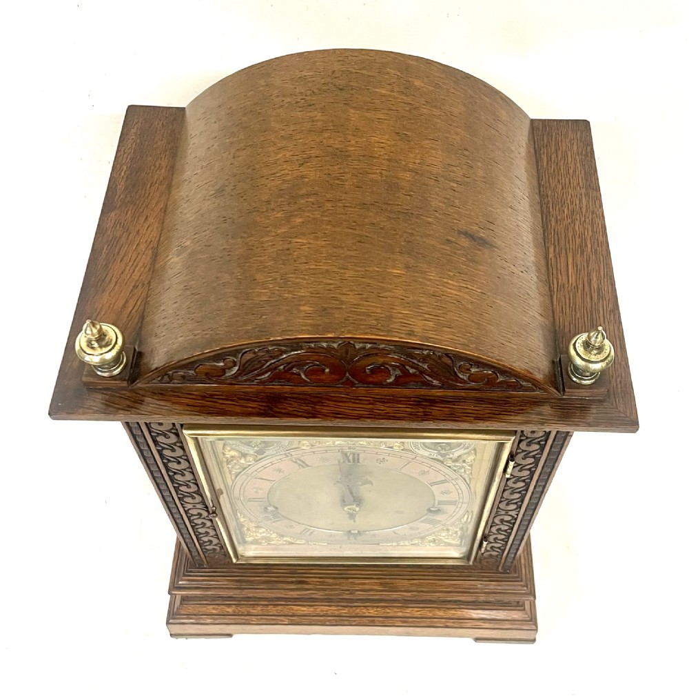 A carved oak bracket clock featuring an arched pediment, brass corner finials, side fretwork panels, and a brass dial with silvered chapter ring. View XV