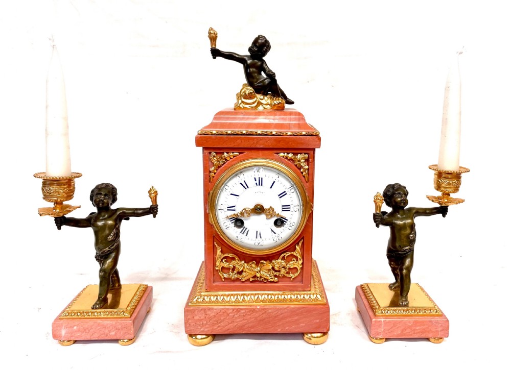 A three-piece antique French garniture set featuring a pink marble and gilt bronze mantel clock flanked by two bronze cherub candelabra.