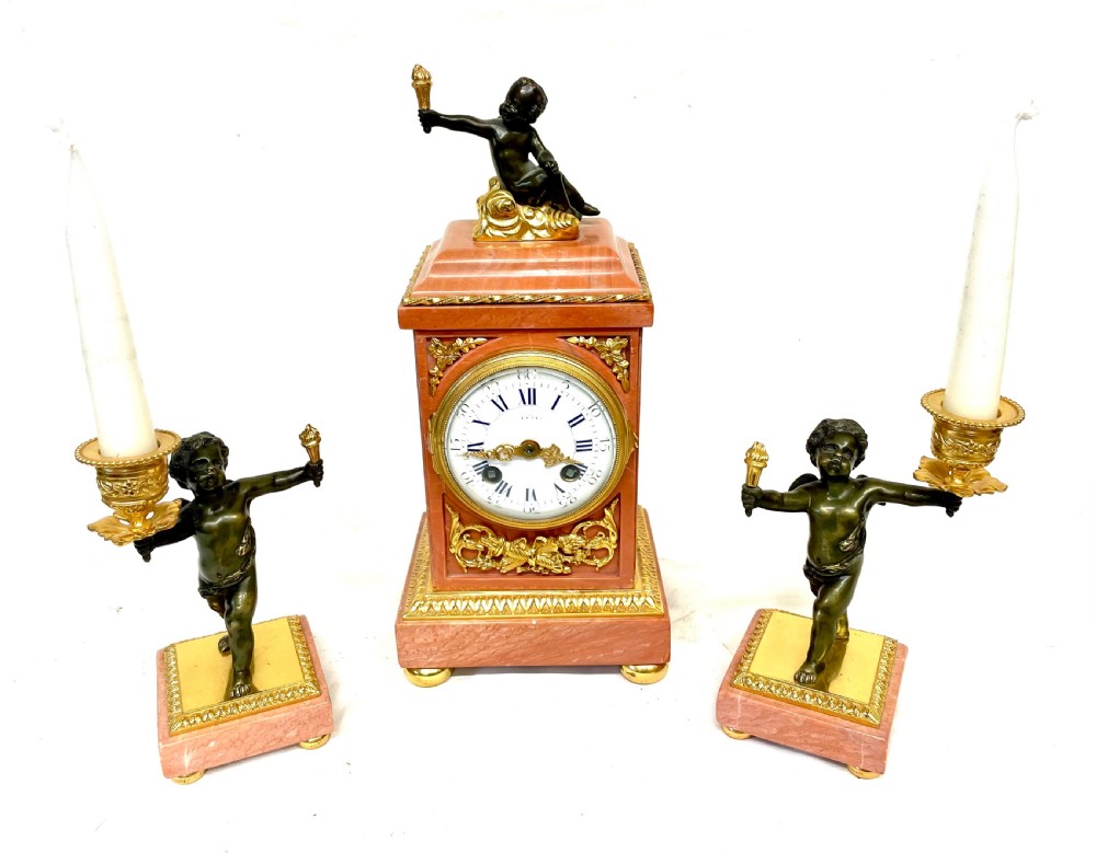 A three-piece antique French garniture set featuring a pink marble and gilt bronze mantel clock flanked by two bronze cherub candelabra. View II