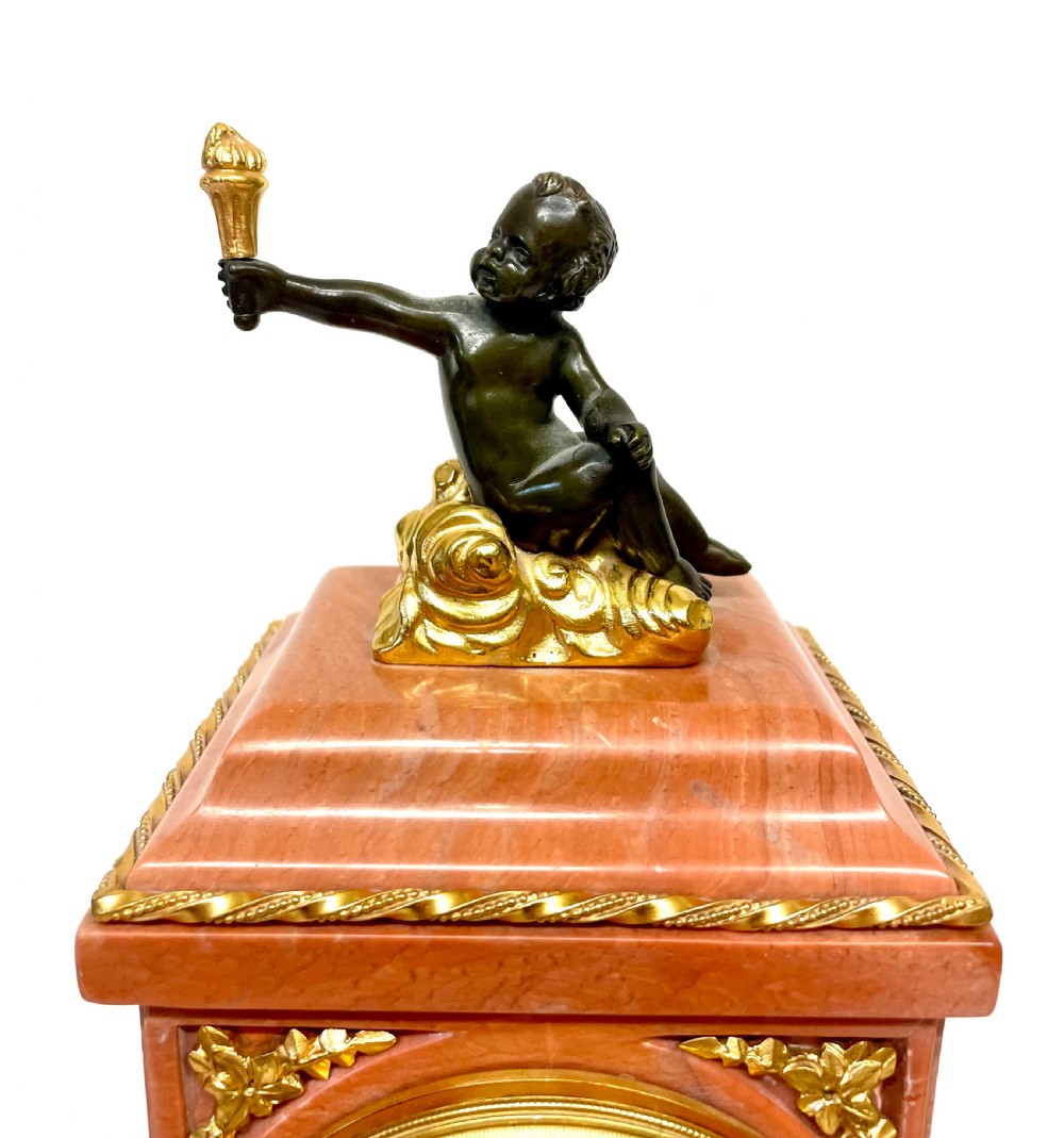 A three-piece antique French garniture set featuring a pink marble and gilt bronze mantel clock flanked by two bronze cherub candelabra. View V